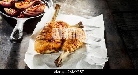 Three grilled spicy seasoned chicken drumsticks served on crumpled white paper with sauteed or roasted vegetables in a skillet Stock Photo