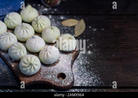 Frozen khinkali on a wooden board, sprinkled with flour. In the background pepper and bay leaf. On a black wooden background. Copy space. Stock Photo