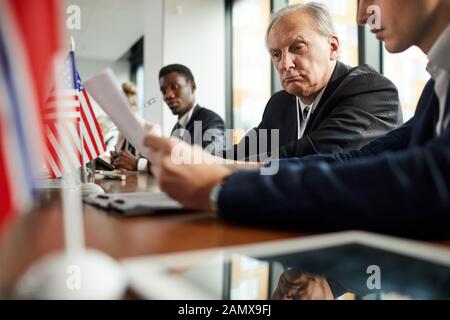 Serious senior political leader sitting at the table with his colleagues and reading the report with them at press conference Stock Photo