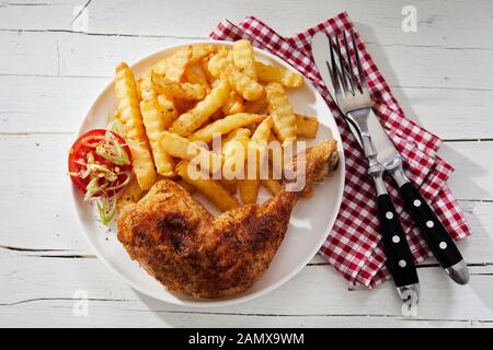 Spicy grilled chicken thigh and leg served on a white plate with crinkle cut oven baked potato chips and salad trimmings viewed top down on rustic whi Stock Photo