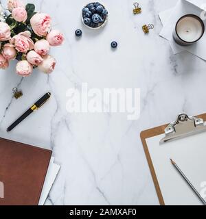 Top view of female home office with copy space in center. Clipboard, flowers, scented candle on white marble. Feminine home office mock up with copy space for text or design. Flat lay Stock Photo