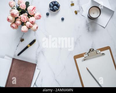 Top view of female home office with copy space in center. Clipboard, flowers, scented candle on white marble. Feminine home office mock up with copy space for text or design. Flat lay Stock Photo