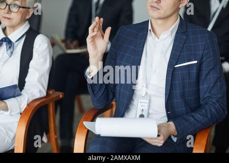 Close-up of businessman in suit sitting with document and raising his hand he taking part at seminar Stock Photo