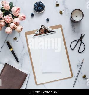 Wedding mock up. Flat lay with blank greeting card, envelope. Clipboard, flowers, scented candle on white marble. Feminine home office mock up with copy space for text or design. Flat lay Stock Photo