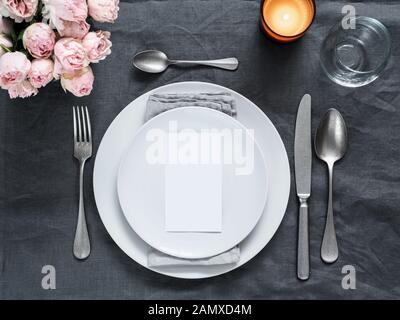 Menu, wedding invitation mock up. Beautiful table setting on gray linen tablecloth. Festive table setting for wedding dinner with pink spray roses and cabdle. Holiday dinner, white plates. Copy space Stock Photo