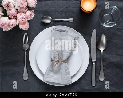 Beautiful table setting with pink spray roses and candle on gray linen tablecloth. Festive table setting for wedding dinner with gray linen napkin on plate. Holiday dinner at home, with white plates Stock Photo