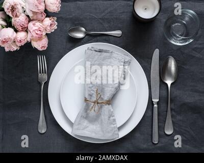 Beautiful table setting with pink spray roses and candle on gray linen tablecloth. Festive table setting for wedding dinner with gray linen napkin on plate. Holiday dinner at home, with white plates Stock Photo