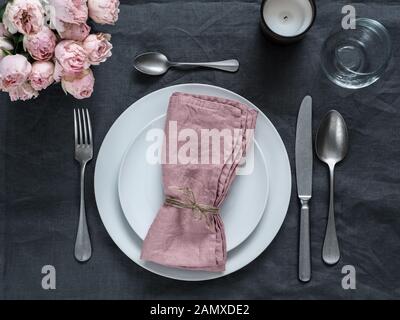 Beautiful table setting with pink spray roses and candle on gray linen tablecloth. Festive table setting for wedding dinner with pink napkin on plate. Holiday dinner with white plates Stock Photo