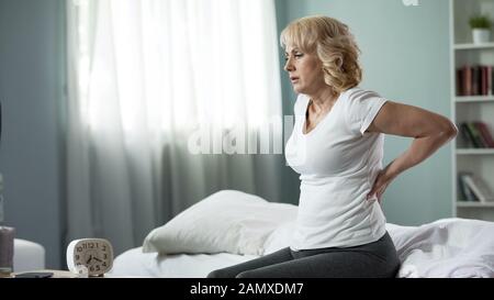 Blond mature lady sitting on bed and touching her back, radiculitis and pain Stock Photo