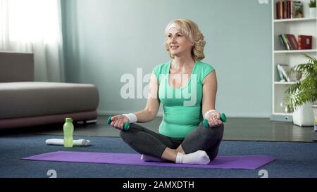 Sportive middle-aged woman holding dumbbells, sitting on mat, home exercises Stock Photo