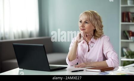 Mature female writer thinking of new series of adventure books in front laptop Stock Photo