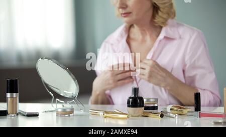 Anti-wrinkles products on table with senior woman looking neck in mirror, beauty Stock Photo