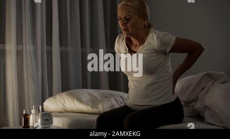 Sick woman touching her back, sitting on bed at night, suffering from pain Stock Photo