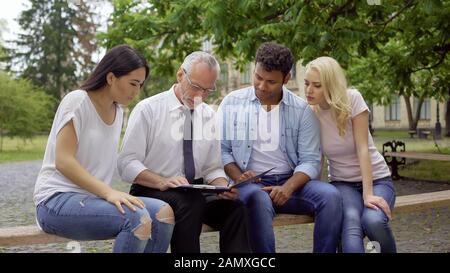 Students and teacher sitting in park near college, discussing science project Stock Photo