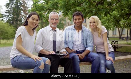 Happy teacher and multi-ethnic students sitting on bench, smiling into camera Stock Photo