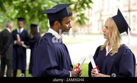 People in academic dress and hats with diplomas in hands talking, graduation day Stock Photo