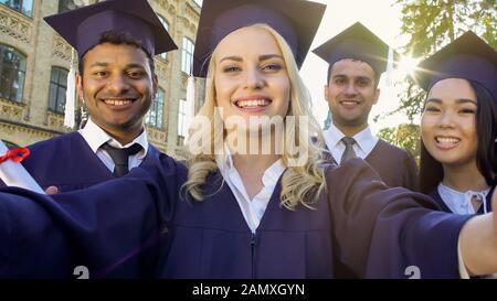 Happy classmates in academic dress taking selfie on graduation day, achievement Stock Photo
