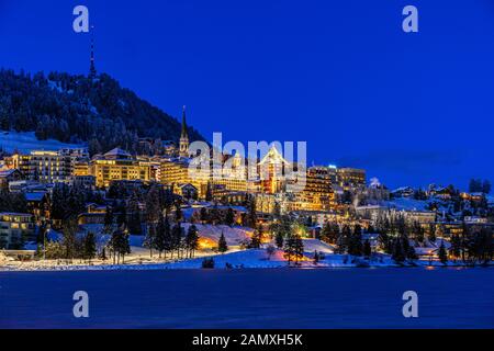 View of beautiful night lights of St. Moritz town in Switzerland at night in winter, with reflection from the lake and snow mountains in backgrouind Stock Photo