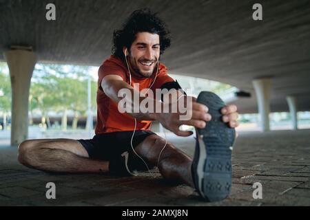 Portrait of a happy man athlete enjoying music on his earphones warming up and stretching his legs before jogging under the bridge Stock Photo