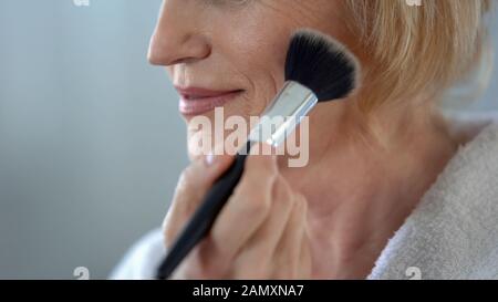 Mature woman applying face powder by make-up brush, preparing for date, beauty Stock Photo