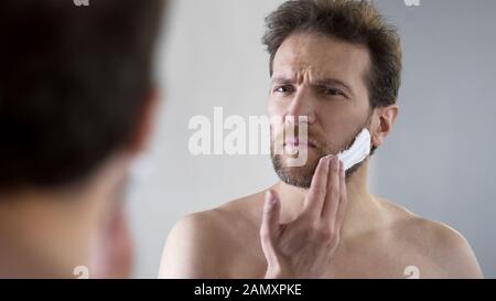 Middle-aged bearded man applying shaving foam on face, morning ritual, mirror Stock Photo