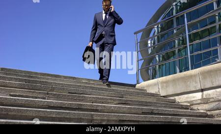 Tired businessman with suitcase talking on phone with partner, going down stairs Stock Photo