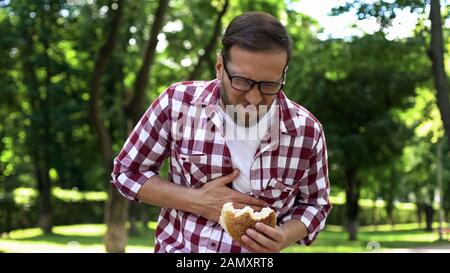 Man with burger in hand feeling nausea, junk food poisoning, body intoxication Stock Photo