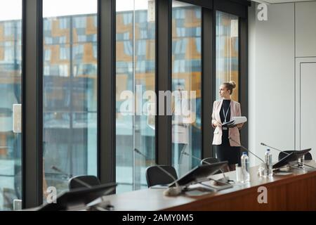 Young female leader holding documents and looking through the window while standing at conference hall Stock Photo