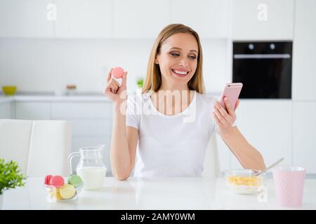 Photo of pretty housewife holding telephone chatting with friends eating colorful macaroons granola breakfast milk on table white light kitchen Stock Photo
