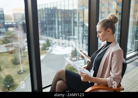 Young businesswoman sitting on chair in front of the big window and examining financial reports Stock Photo