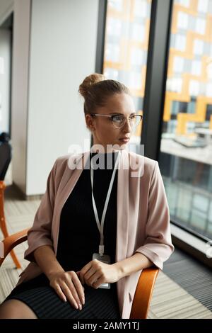Young serious businesswoman in eyeglasses sitting on chair and thinking over new ideas during meeting at office Stock Photo