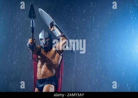 Front view of fearless muscular spartan hiding from rain under shield. Portrait of ancient warrior in iron helmet and red cloak pointing spear in bad rainy evening weather. Concept of warrior, sparta. Stock Photo