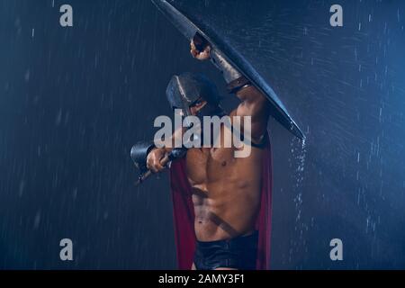 Front view of fearless muscular spartan hiding from rain under shield and looking at camera. Portrait of ancient warrior in iron helmet and red cloak pointing spear in bad rainy evening weather. Stock Photo