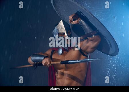 Front view of fearless muscular spartan hiding from rain under shield. Portrait of ancient warrior in iron helmet and red cloak pointing spear in bad rainy evening weather. Concept of warrior, sparta. Stock Photo