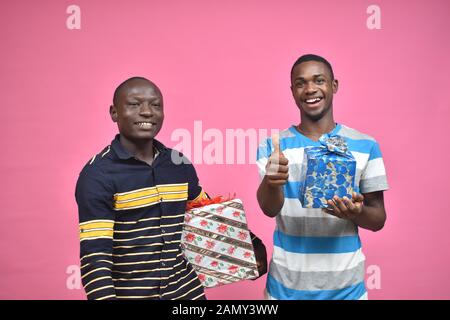 two young black guys holding gift boxes and feeling excited, one giving a thumbs up Stock Photo