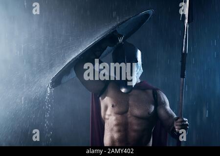 Front view of fearless muscular spartan hiding from rain with shield. Portrait of ancient warrior in iron helmet and red cloak holding spear and looking at camera in bad evening weather. Stock Photo