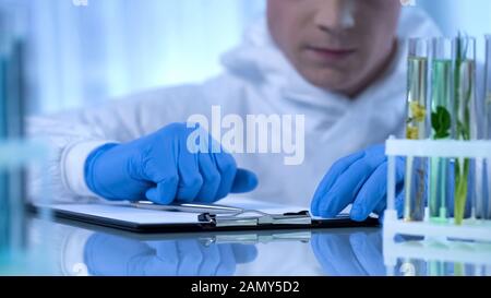 Biologist observing plant in test tube, writing information about growth changes Stock Photo
