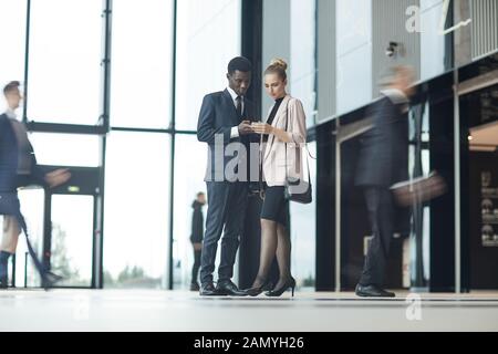 Two young colleagues meeting each other in the hall of office building and using their phones Stock Photo