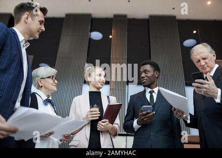 Multiethnic group of business people holding documents and gadgets standing in the office hall and talking to each other Stock Photo