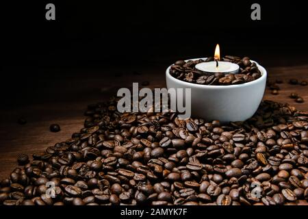 White coffee cup full of coffee beans and burning candle on top of coffee bean stack - image Stock Photo