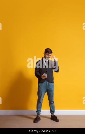 Disguasted. Young caucasian man using smartphone, serfing, chatting, betting. Full length portrait isolated on yellow background. Concept of modern technologies, millennials, social media. Stock Photo