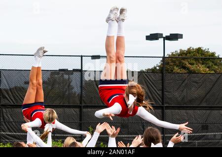 Two high school teenage cheerleaders in red and white uniforms are being flipped in the air with their teammates ready to catch them while practicing Stock Photo