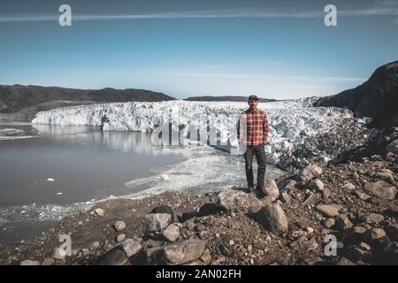 A young man traveler tourist standing in front of Eqip Sermia glacier called Eqi Glacier. Wall of ice in background. The concept of global warming and Stock Photo