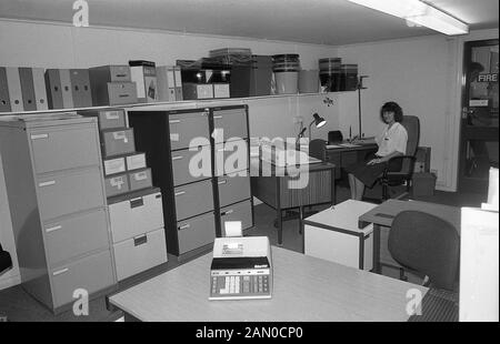 1986, historical, a female clerical worker at a desk in an low-ceiling office, surrounded office supplies and stacks of metal filing cabinets, England, UK. Stock Photo