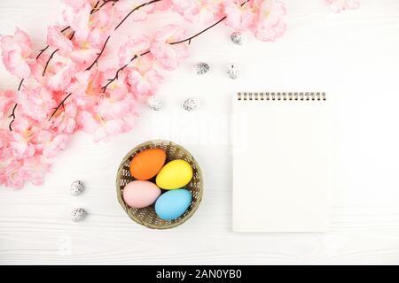 Pastel colored eggs in the nest with the branch of pink flowers on white wooden background. Empty notebook and basket with eggs on Easter background. Stock Photo