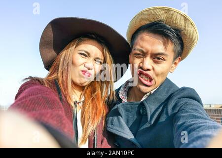 Hipster asian couple making selfie with funny faces - Lovers having fun together outdoor - Technology and fashion lifestyle concept - Soft brown vinta Stock Photo