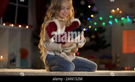 Adorable girl playing with teddy bear under glowing Christmas tree, holiday eve Stock Photo