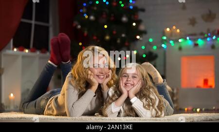 Young woman and girl lying under Christmas tree, smiling to camera, holidays Stock Photo
