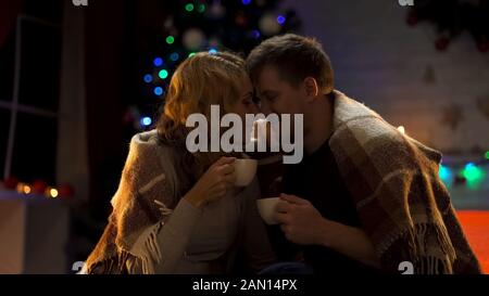 Loving wife and husband drinking hot cocoa under cozy plaid and nuzzling, X-mas Stock Photo