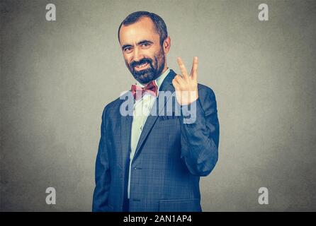 Portrait headshot middle aged businessman smiling with victory hand sign symbol isolated grey wall office background. Positive emotion face expression Stock Photo